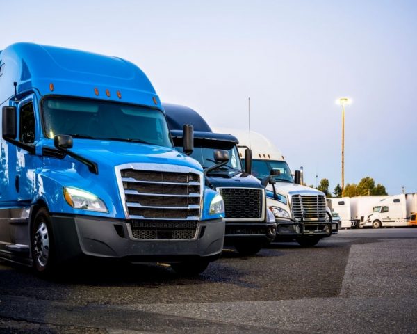 Different makes and models industrial grade professional big rigs semi trucks standing in row on the truck stop parking lot at evening time waiting to continued delivery schedule