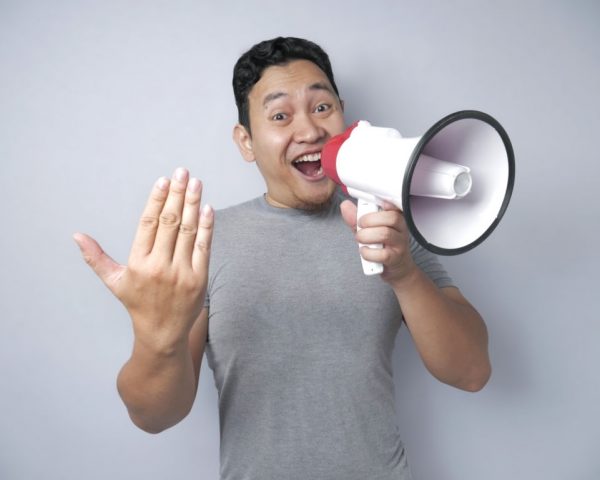 Young Asian man making announcement, advertisement concept, Smiling expression using megaphone. Close up body portrait over grey background