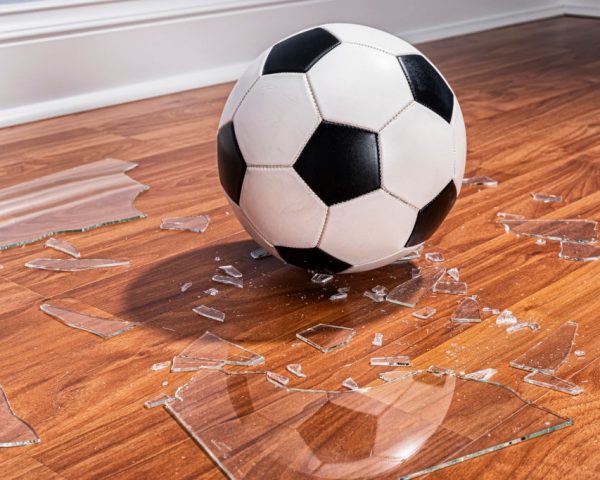 A soccer ball sitting among pieces of a broken window glass that are on the wood floor inside a home.